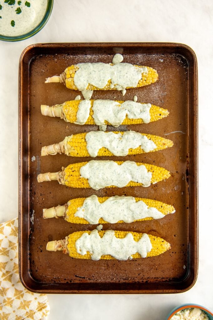 Six ears of corn on the cob are arranged in a row on a baking sheet, each topped with a generous drizzle of cilantro lime crema sauce. A patterned napkin and small bowls are partially visible nearby.
