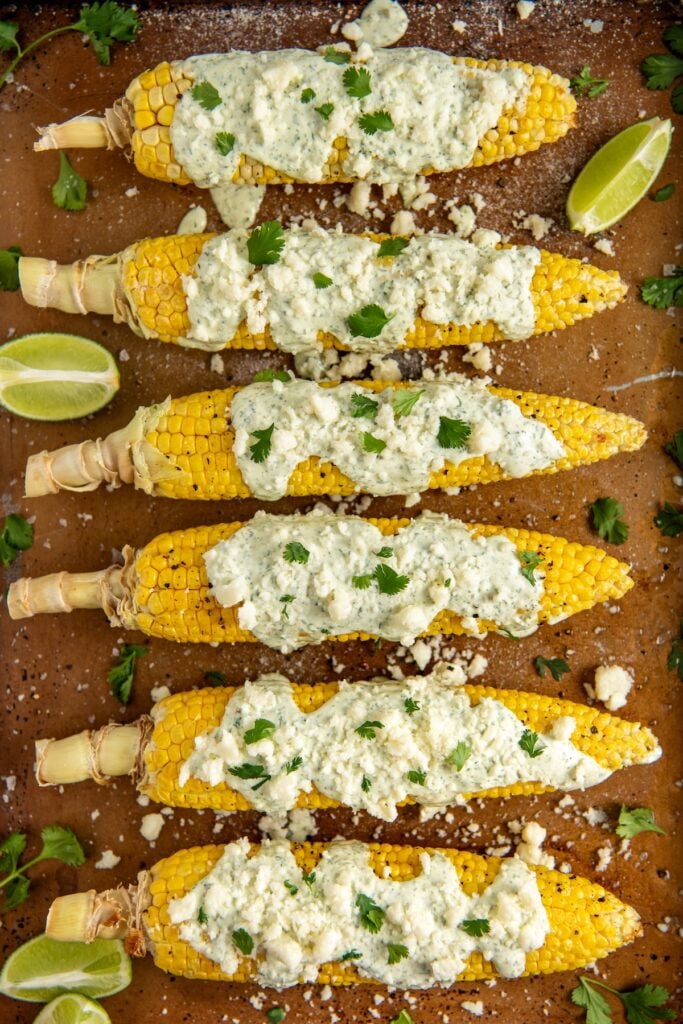 Six ears of creamy cilantro lime corn on the cob are laid out on a tray, topped with cilantro lime crema sauce, crumbled cheese, and fresh cilantro. Lime wedges and scattered herbs are also visible on the tray.