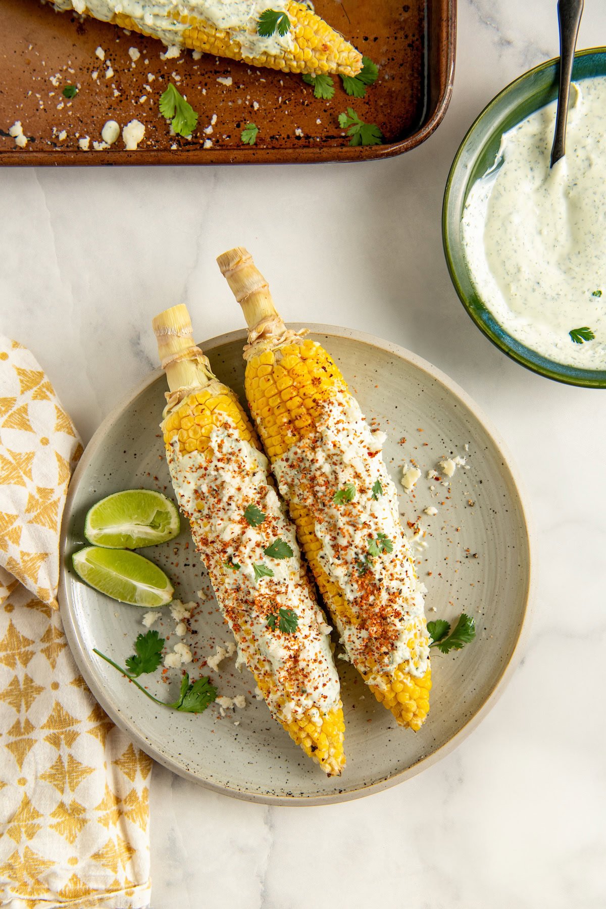 Two ears of cilantro street corn on the cob, topped with white sauce, cheese, and chili powder, are served on a plate with lime wedges and cilantro, next to a bowl of creamy sauce and a yellow-patterned napkin on a marble surface.