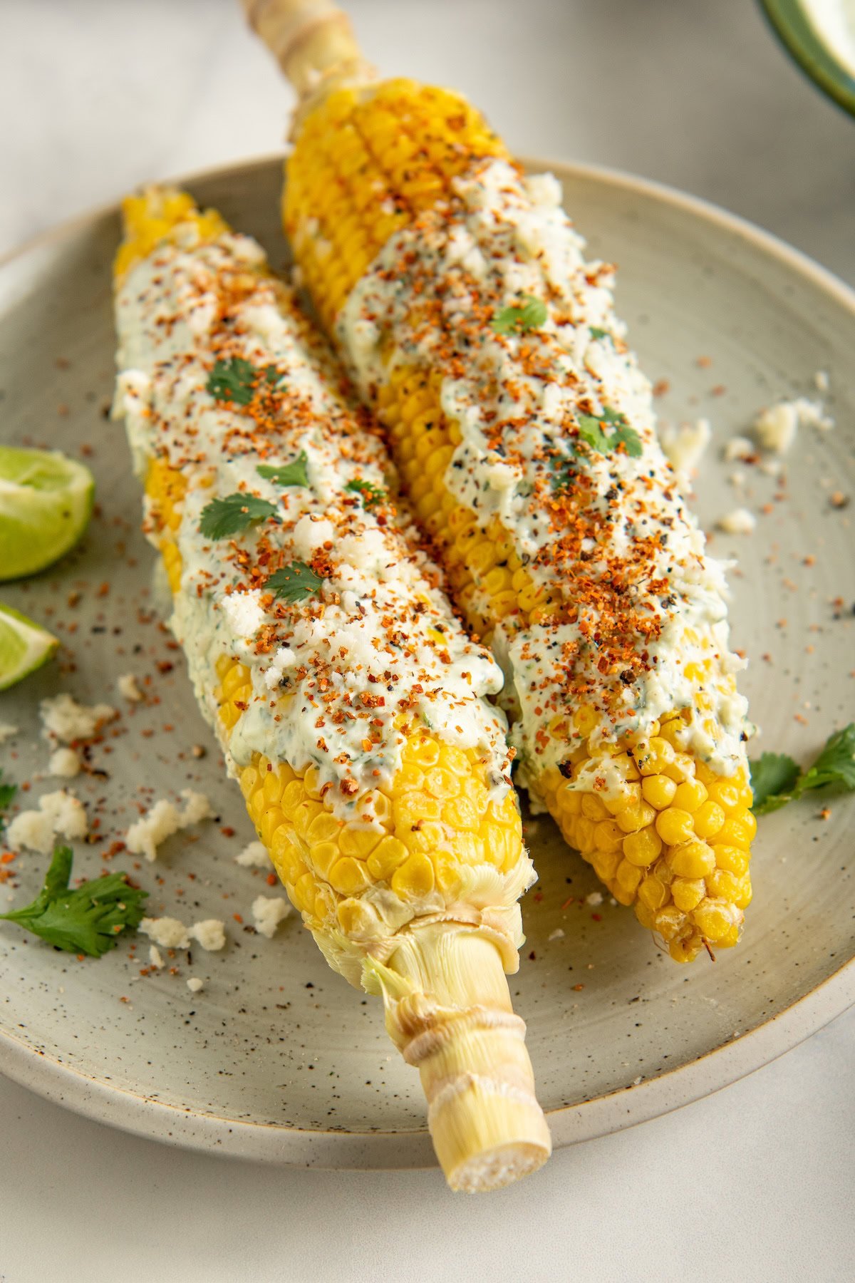 Two ears of cilantro lime corn on the cob topped with creamy sauce, crumbled cheese, chili powder, and cilantro on a plate, with lime wedges on the side.