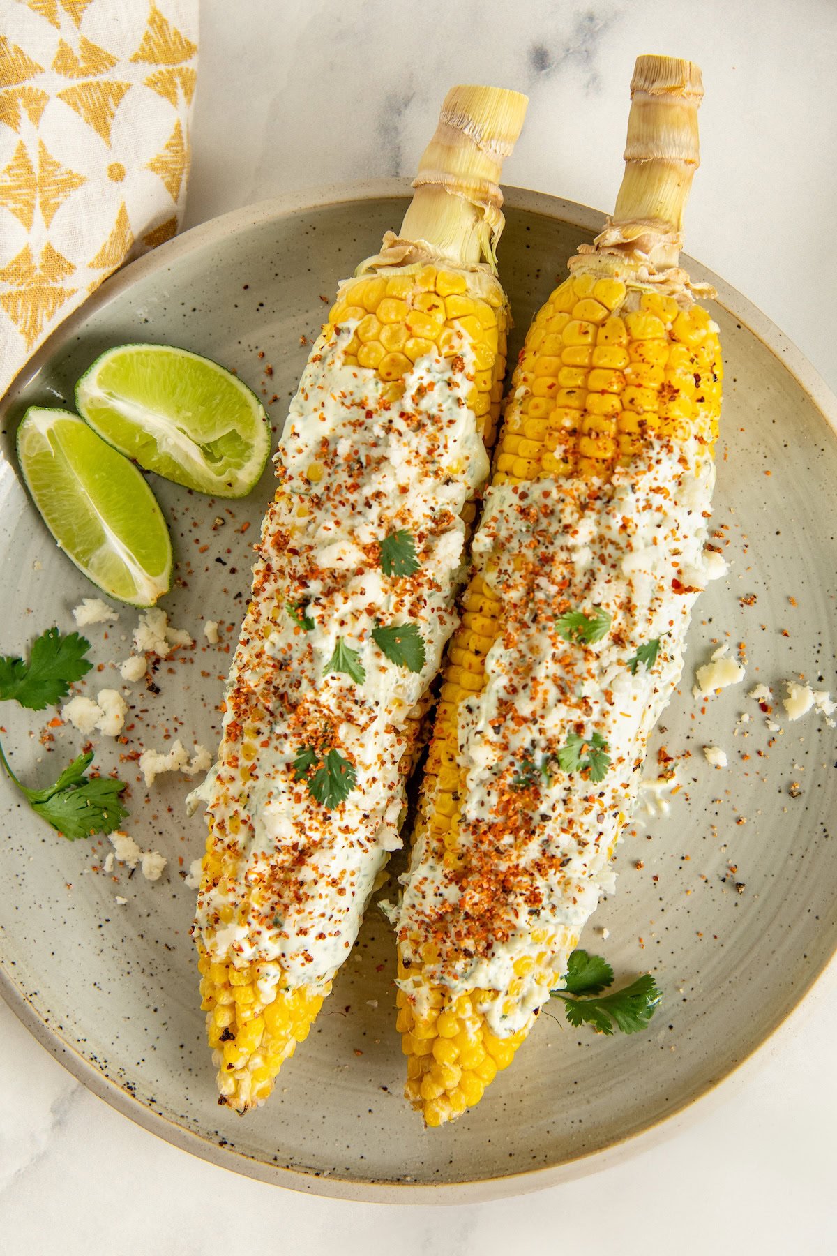 Two ears of cilantro lime corn on the cob covered in cilantro lime crema, sprinkled with crumbled cheese, tan, and cilantro, served on a plate with lime wedges and a few cilantro leaves.