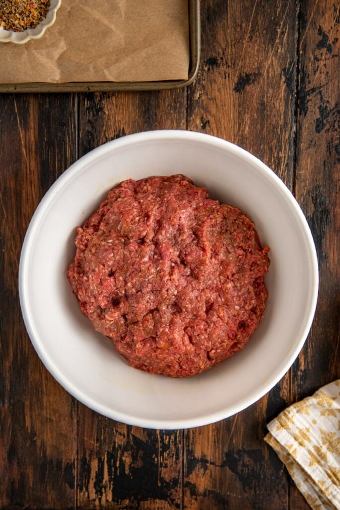 A white bowl filled with raw ground meat sits on a wooden table. A small dish with spices and a baking tray are partially visible in the background alongside a patterned cloth.