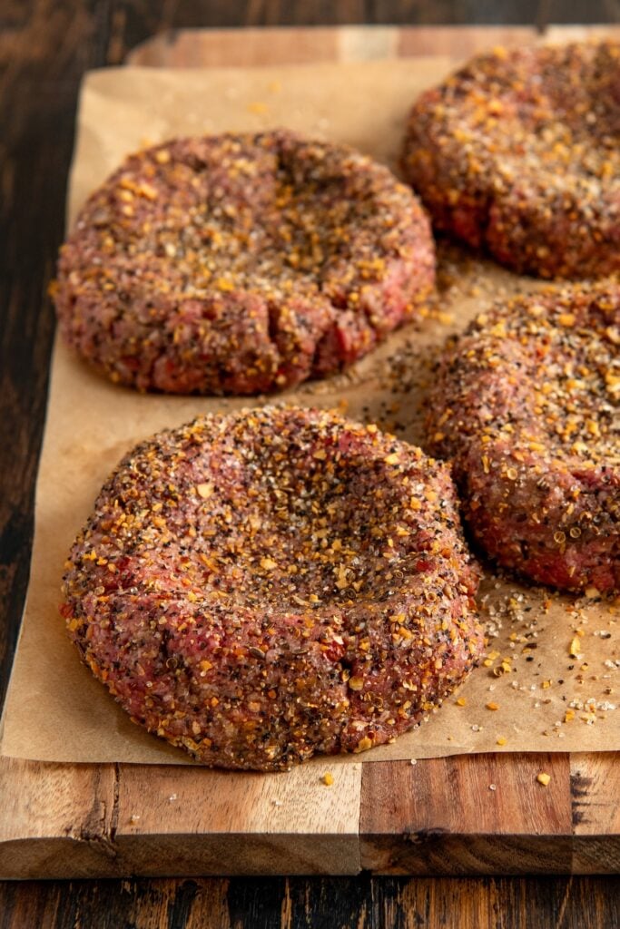 Four raw, seasoned beef burger patties with a dimple in the center rest on parchment paper atop a wooden cutting board.