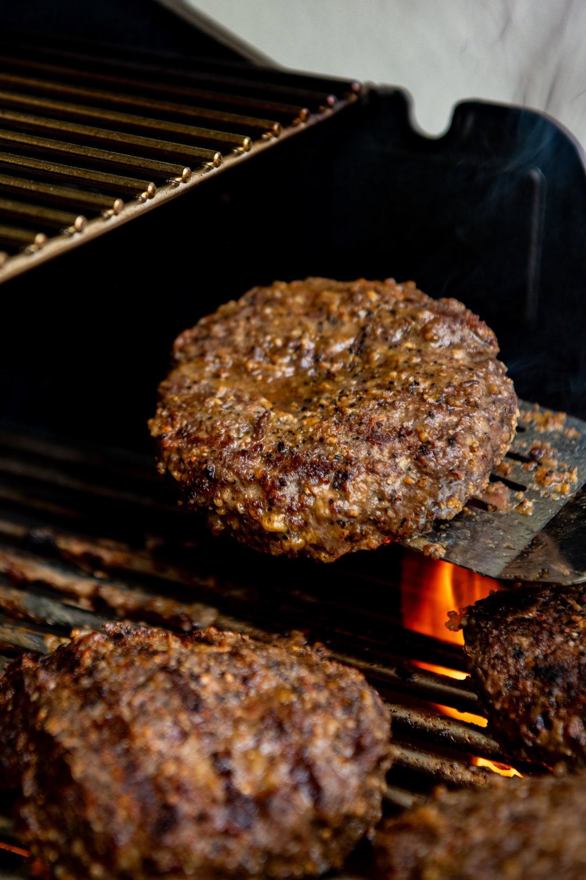 A close-up of a grilled hamburger patty being lifted off a hot grill with flames underneath, while other patties cook beside it.