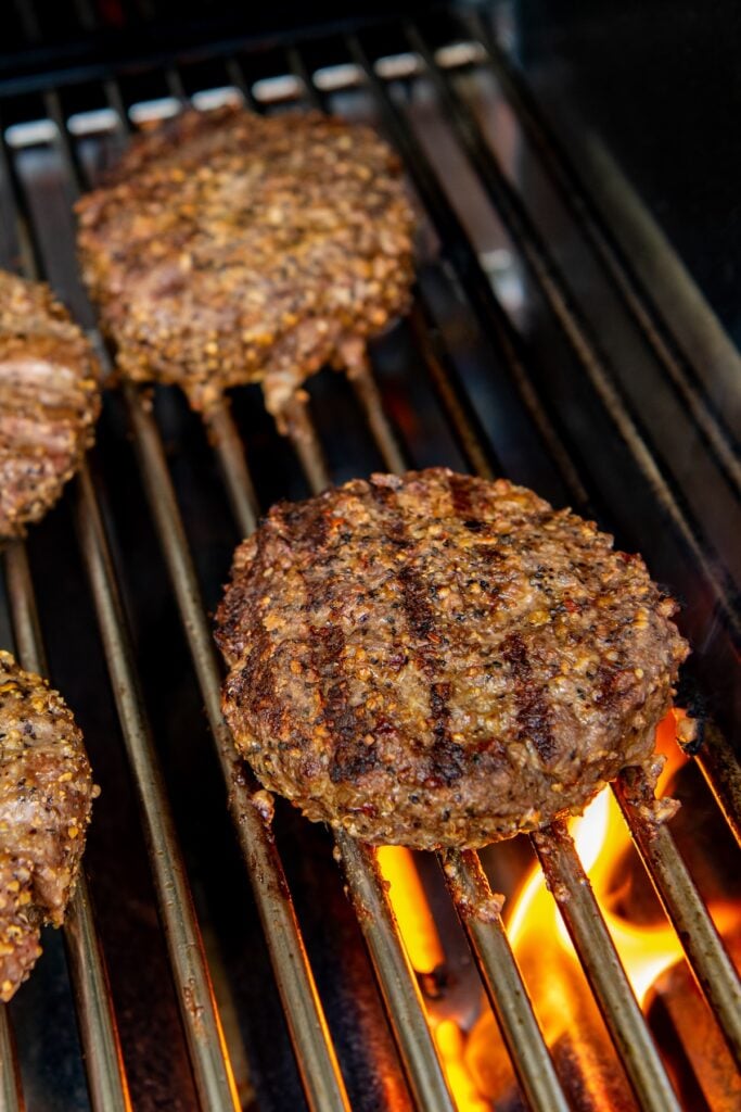 Four beef burger patties cooking on a grill, with visible grill marks and an open flame beneath them. The patties are browned and sizzling over the heat.