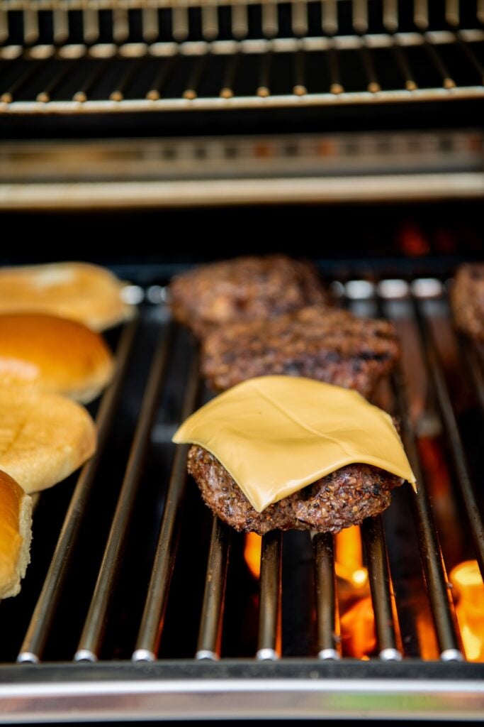 A cheeseburger patty with melted cheddar cheese grills over an open flame next to burger buns and other patties on a barbecue grill.