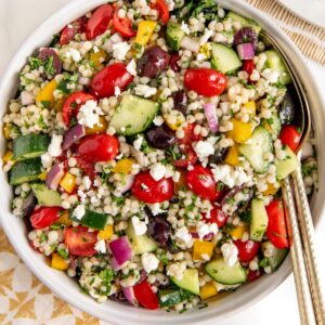 A bowl of Mediterranean couscous salad with cherry tomatoes, cucumbers, red onions, yellow bell peppers, kalamata olives, feta cheese, and fresh herbs, with a serving spoon on the side.
