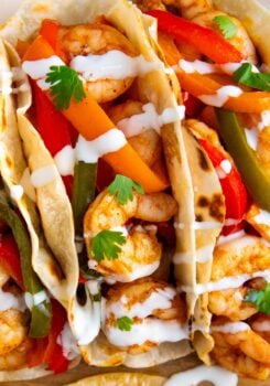 Close-up of shrimp tacos filled with sautéed shrimp, sliced red and green bell peppers, and carrots, topped with fresh cilantro leaves and drizzled with a white sauce in soft, lightly toasted tortillas.