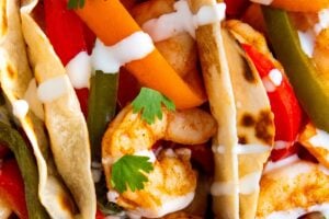Close-up of shrimp tacos filled with sautéed shrimp, sliced red and green bell peppers, and carrots, topped with fresh cilantro leaves and drizzled with a white sauce in soft, lightly toasted tortillas.