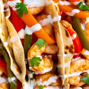 Close-up of shrimp tacos filled with sautéed shrimp, sliced red and green bell peppers, and carrots, topped with fresh cilantro leaves and drizzled with a white sauce in soft, lightly toasted tortillas.