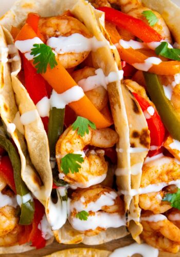 Close-up of shrimp tacos filled with sautéed shrimp, sliced red and green bell peppers, and carrots, topped with fresh cilantro leaves and drizzled with a white sauce in soft, lightly toasted tortillas.