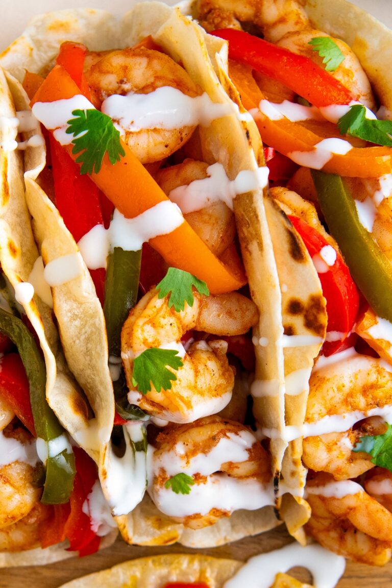 Close-up of shrimp tacos filled with sautéed shrimp, sliced red and green bell peppers, and carrots, topped with fresh cilantro leaves and drizzled with a white sauce in soft, lightly toasted tortillas.