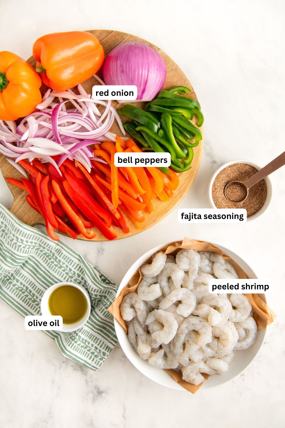 Labels mark each ingredient for this sheet pan shrimp fajita recipe. A round cutting board with sliced red onion, green, orange, and red bell peppers, a bowl of fajita seasoning with a spoon, a bowl of peeled shrimp, and a small bowl of olive oil on a striped towel.
