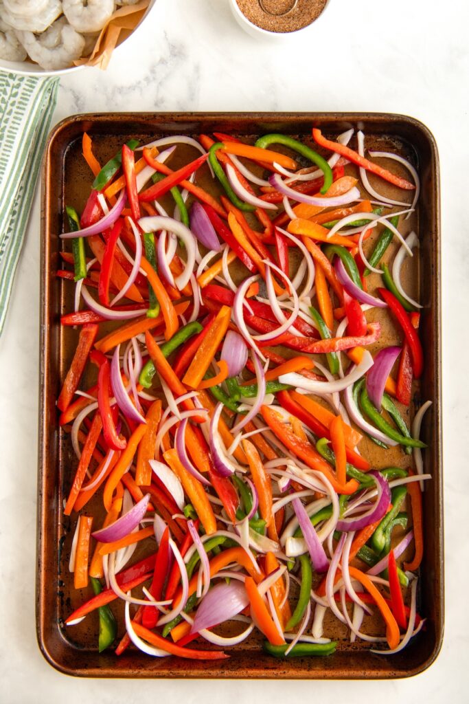 A baking sheet filled with sliced red, orange, and green bell peppers and onions, ready to be roasted. Striped kitchen towel, bowl of seasoning, and a basket with shrimp are visible nearby.