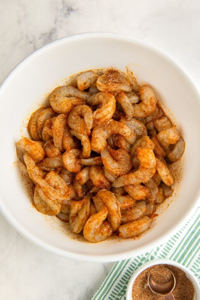 Raw shrimp coated with a fajita seasoning mix in a white bowl, sitting on a white surface next to a green-striped cloth and a small metal measuring cup with extra seasoning.
