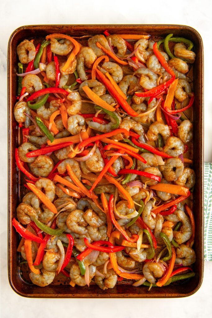 A sheet pan filled with raw shrimp, sliced red, orange, and green bell peppers, and onion strips, all seasoned and ready to be cooked.