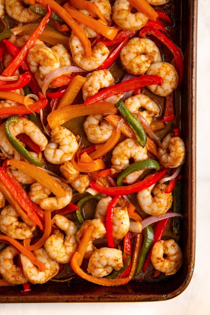 Sheet pan with cooked shrimp, sliced red, yellow, and green bell peppers, and red onion strips, all seasoned and roasted together.
