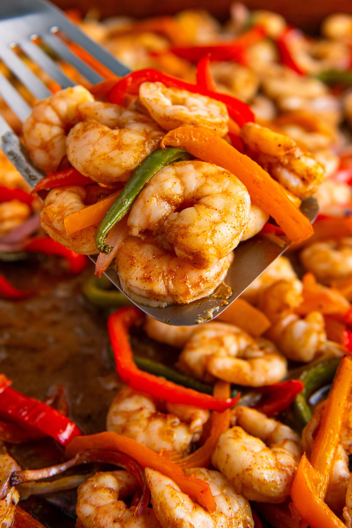 A spatula lifts seasoned, cooked shrimp fajitas mixed with sliced red, orange, and green bell peppers off a sheet pan.