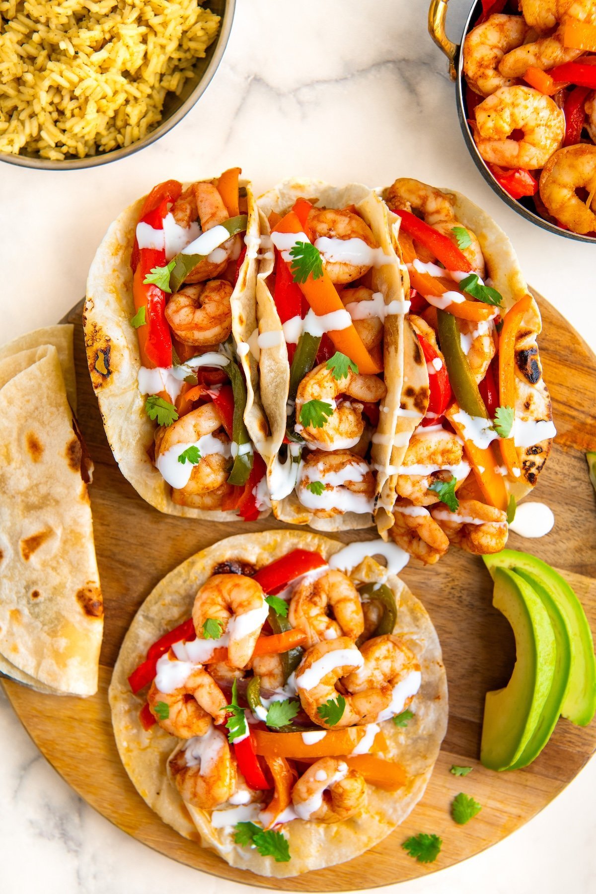 Three shrimp fajita tacos served in flour tortillas with colorful bell peppers, drizzled with white sauce and garnished with cilantro, served on a wooden board with avocado slices, rice, shrimp, and extra tortillas nearby.