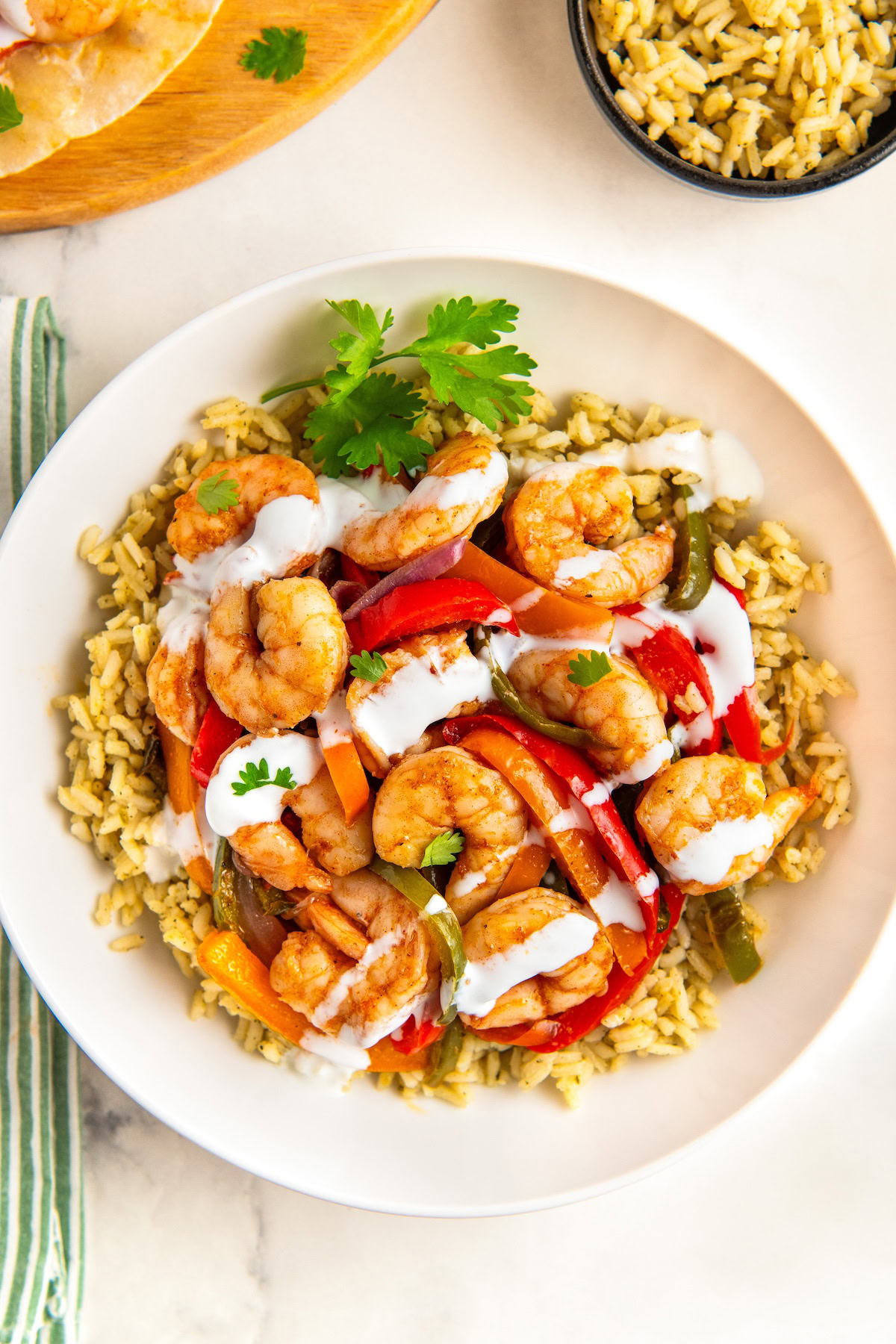 A white plate filled with sheet pan shrimp fajitas with sautéed bell peppers and onions on a bed of seasoned rice, topped with a drizzle of sour cream and garnished with cilantro. A bowl of rice is visible nearby.