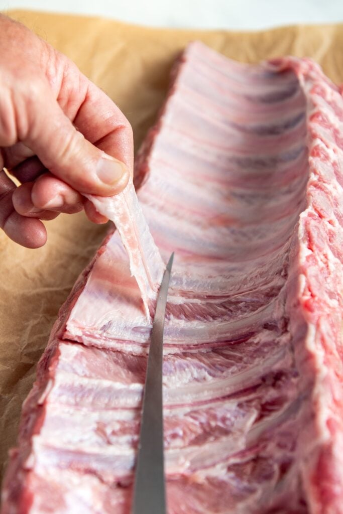 A hand uses a knife to remove the thin membrane from the back of a raw rack of pork ribs placed on brown parchment paper.