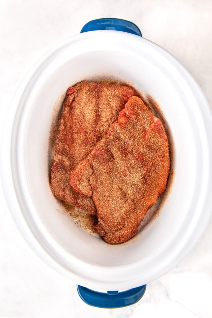 Two raw pork ribs coated with a layer of dry seasoning are placed inside a white slow cooker with a blue handle, viewed from above on a light-colored surface.