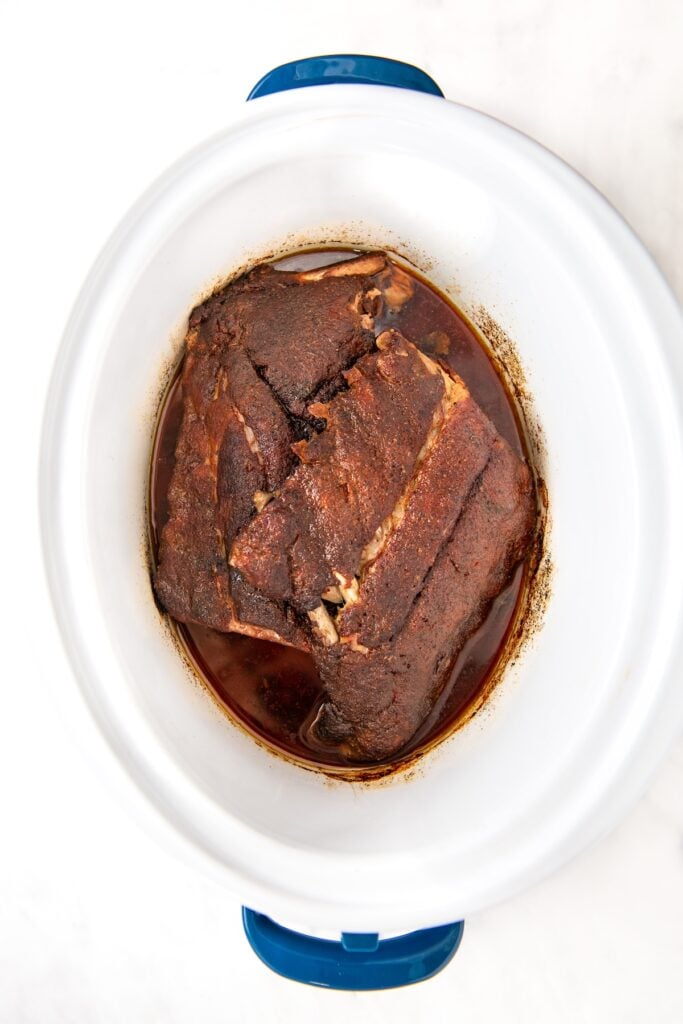 Cooked and seasoned pork ribs sits in liquid from cooking inside a white slow cooker.
