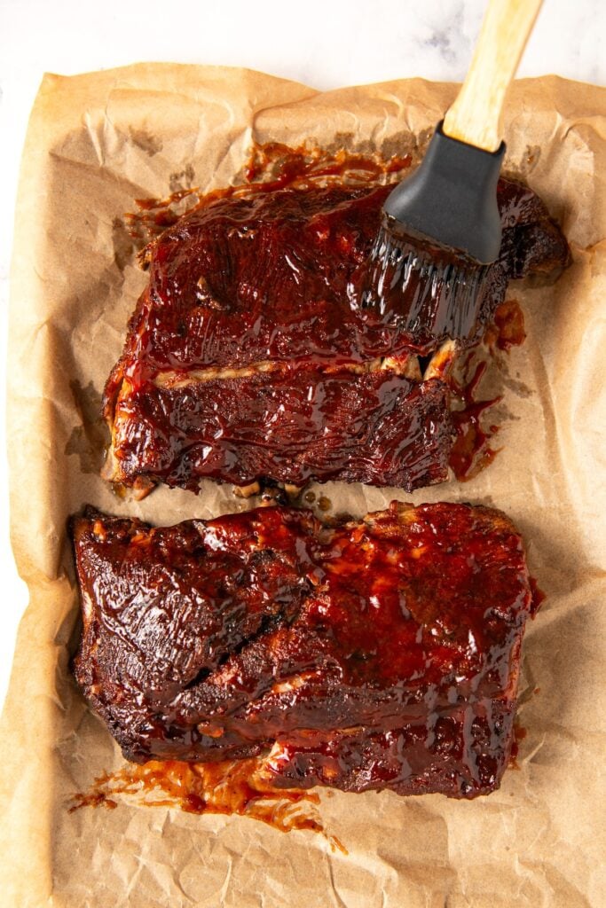 Two racks of glazed barbecue pork ribs on parchment paper, with a brush applying sauce to the top rack.