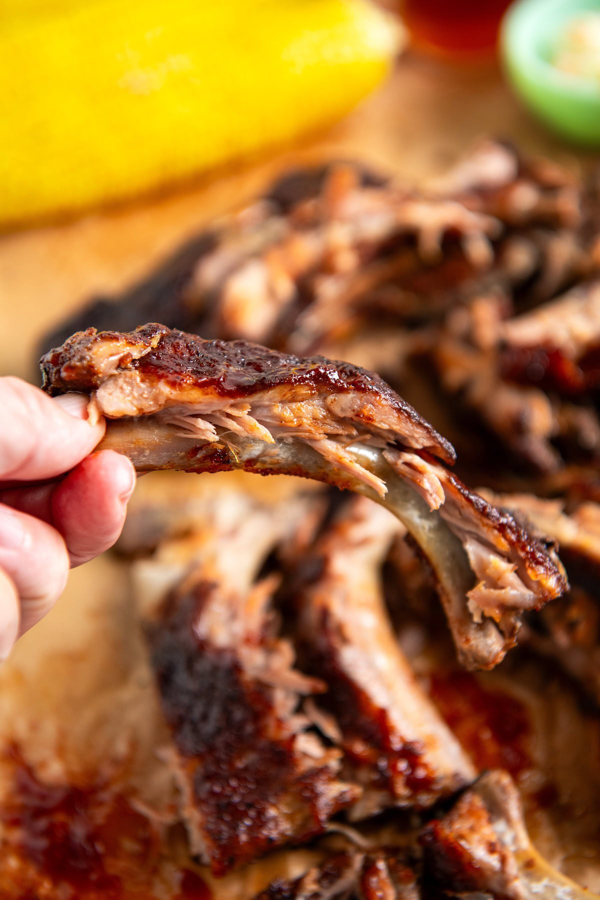 A hand holding a tender, saucy slow cooker baby back rib with a bite taken out, in front of a pile of pork ribs and a blurred corn on the cob in the background.