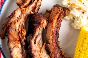 A plate with three slow cooker baby back ribs, a portion of creamy coleslaw, and a piece of corn on the cob, set on a red and white checkered tablecloth.
