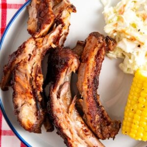 A plate with three slow cooker baby back ribs, a portion of creamy coleslaw, and a piece of corn on the cob, set on a red and white checkered tablecloth.