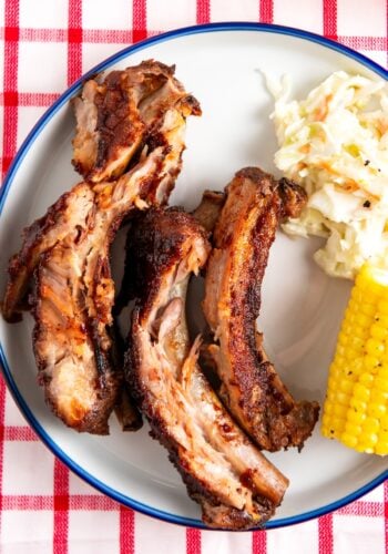 A plate with three slow cooker baby back ribs, a portion of creamy coleslaw, and a piece of corn on the cob, set on a red and white checkered tablecloth.