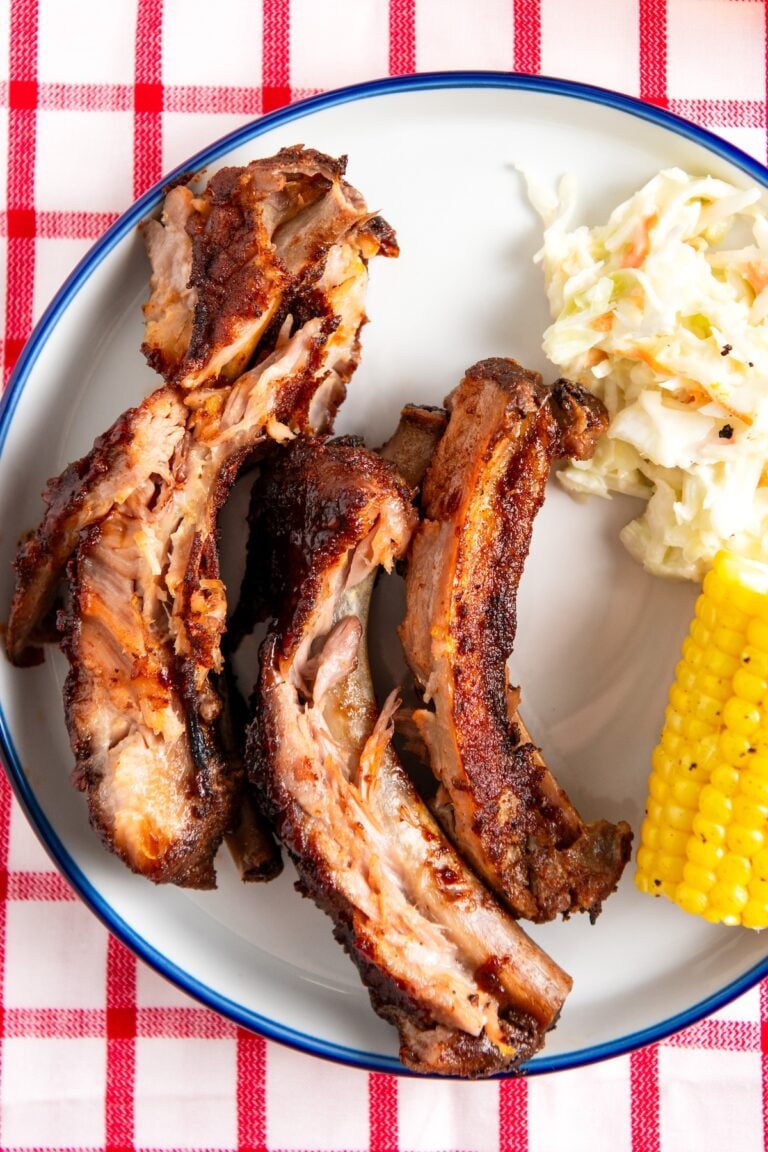 A plate with three slow cooker baby back ribs, a portion of creamy coleslaw, and a piece of corn on the cob, set on a red and white checkered tablecloth.