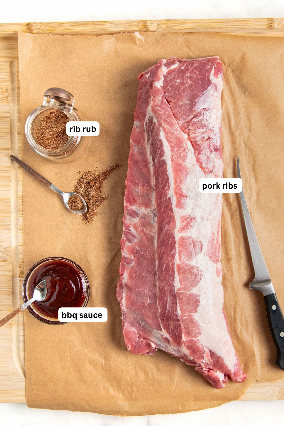A slab of raw pork baby back ribs on parchment paper, with a jar of rib rub, a spoonful of seasoning, a jar of BBQ sauce, and a knife, all labeled. The setup is on a wooden cutting board.