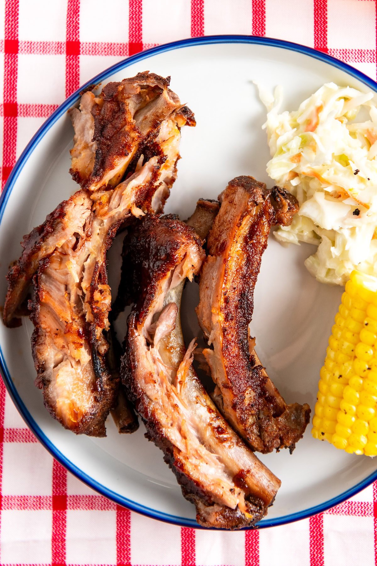 A plate with three slow cooker baby back ribs, a portion of creamy coleslaw, and a piece of corn on the cob, set on a red and white checkered tablecloth.