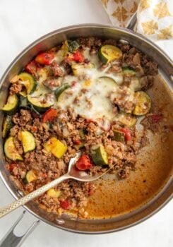 A skillet filled with cooked ground beef and zucchini, with tomatoes, onions, and melted cheese on top. A metal serving spoon is scooping from the mixture.