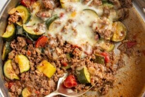 A skillet filled with cooked ground beef and zucchini, with tomatoes, onions, and melted cheese on top. A metal serving spoon is scooping from the mixture.