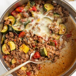 A skillet filled with cooked ground beef and zucchini, with tomatoes, onions, and melted cheese on top. A metal serving spoon is scooping from the mixture.