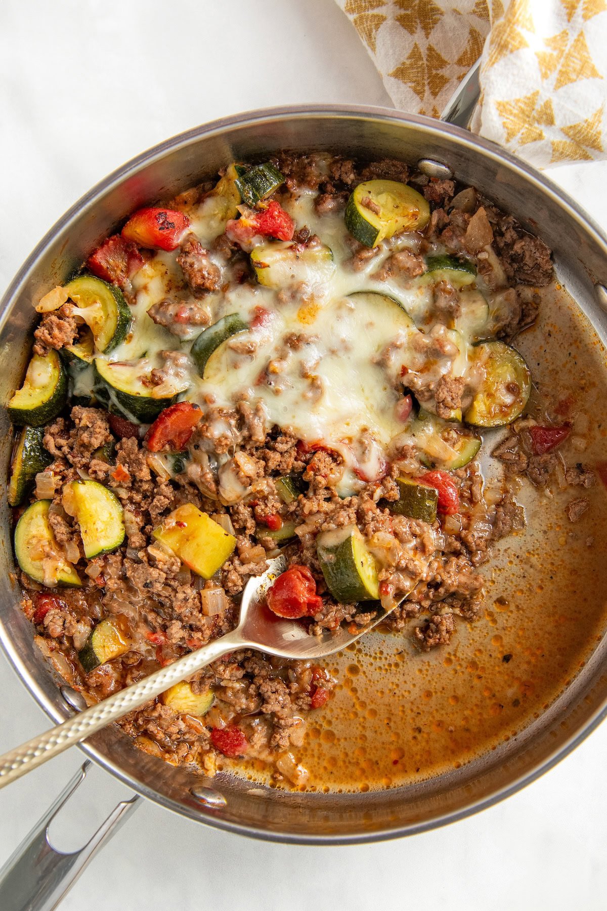 Ready in 30 minutes, this low-carb Cheesy Ground Beef Zucchini Skillet is boldly seasoned and topped with lots of gooey mozzarella cheese. A skillet filled with cooked ground beef and zucchini, with tomatoes, onions, and melted cheese on top. A metal serving spoon is scooping from the mixture.