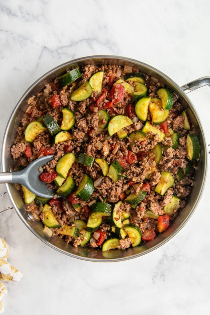 Ready in 30 minutes, this low-carb Cheesy Ground Beef Zucchini Skillet is boldly seasoned and topped with lots of gooey mozzarella cheese. A skillet filled with a cooked mixture of ground beef, zucchini slices, diced tomatoes, and onions, with a serving spoon resting inside, all on a marble countertop.