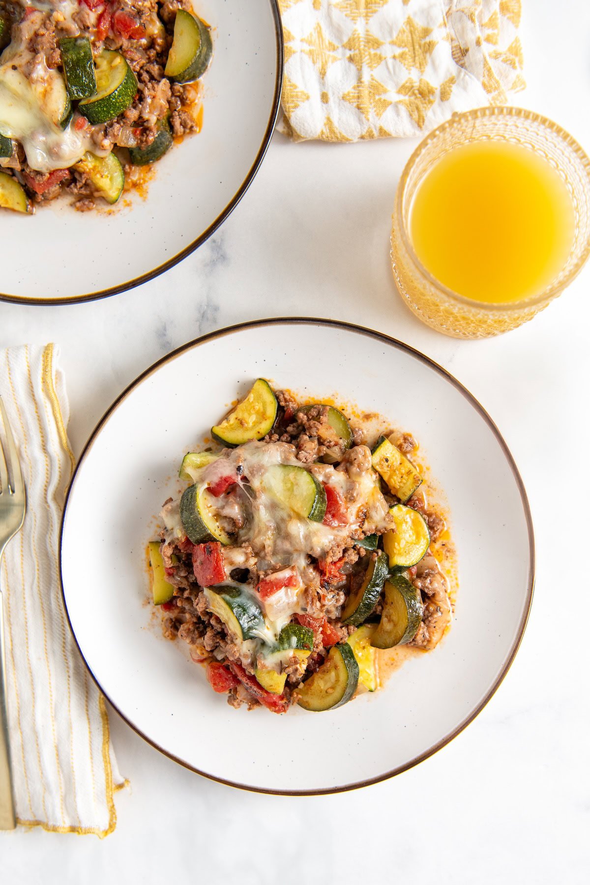 Ready in 30 minutes, this low-carb Cheesy Ground Beef Zucchini Skillet is boldly seasoned and topped with lots of gooey mozzarella cheese. A plate of cheesy ground beef and zucchini with tomatoes and melted cheese on top. Served with a glass of juice and a patterned napkin on a white surface.