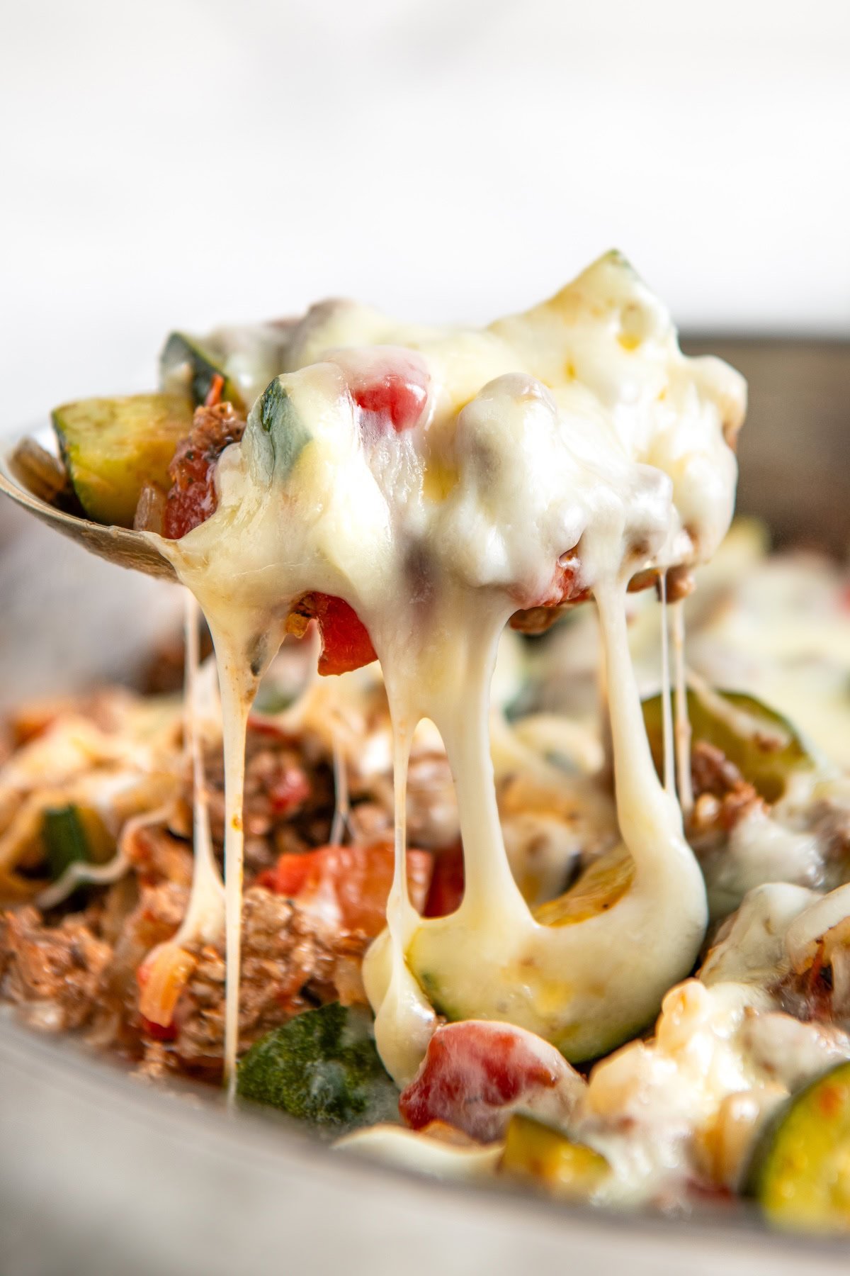 Ready in 30 minutes, this low-carb Cheesy Ground Beef Zucchini Skillet is boldly seasoned and topped with lots of gooey mozzarella cheese. A spoon lifting a cheesy zucchini and ground beef skillet dinner, with melted cheese stretching and chunks of zucchini, tomato, and ground meat visible.