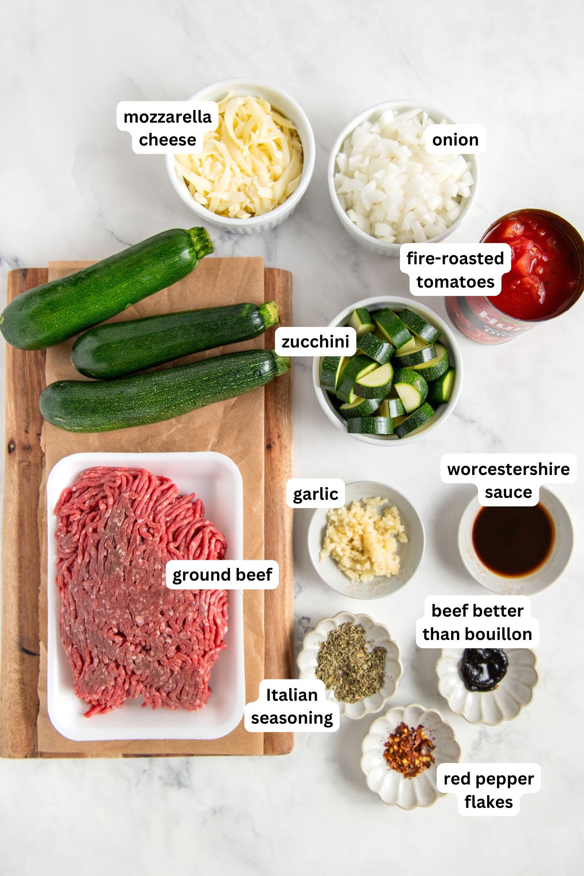 Ready in 30 minutes, this low-carb Cheesy Ground Beef Zucchini Skillet is boldly seasoned and topped with lots of gooey mozzarella cheese. Top-down view of labeled ingredients on a white surface, including mozzarella cheese, chopped onion, fire-roasted tomatoes, whole and sliced zucchini, ground beef, minced garlic, Worcestershire sauce, beef bouillon, Italian seasoning, and red pepper flakes.