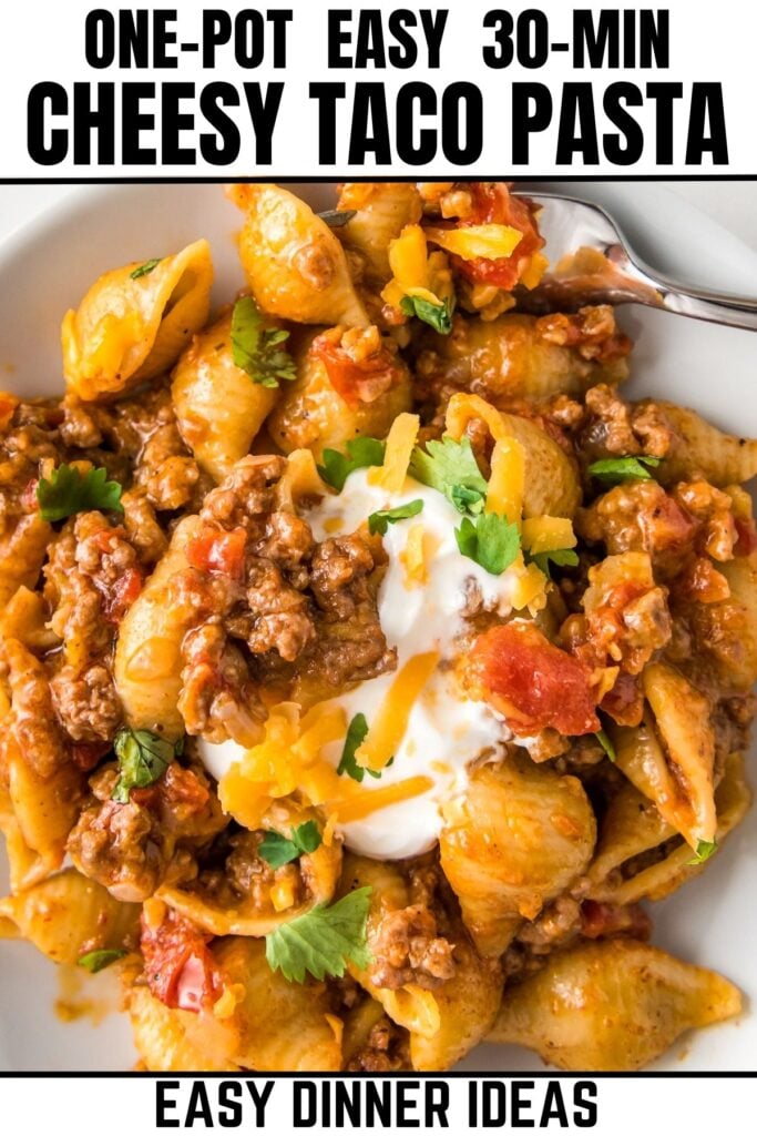 A plate of cheesy taco pasta made with shell pasta, ground meat, diced tomatoes, melted cheese, and topped with sour cream, shredded cheese, and cilantro.