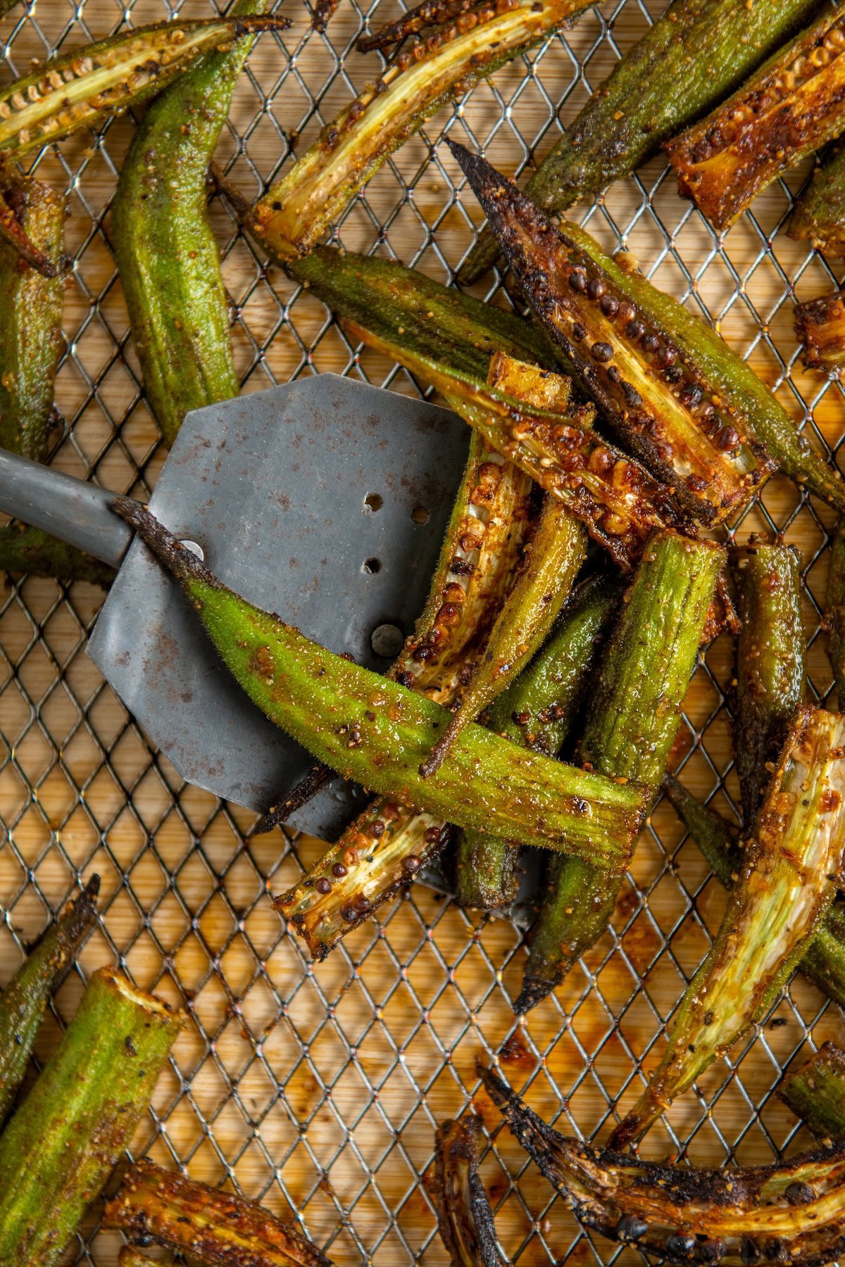 A metal spatula lifts charred okra spears from a wire rack, showing browned and crispy edges.