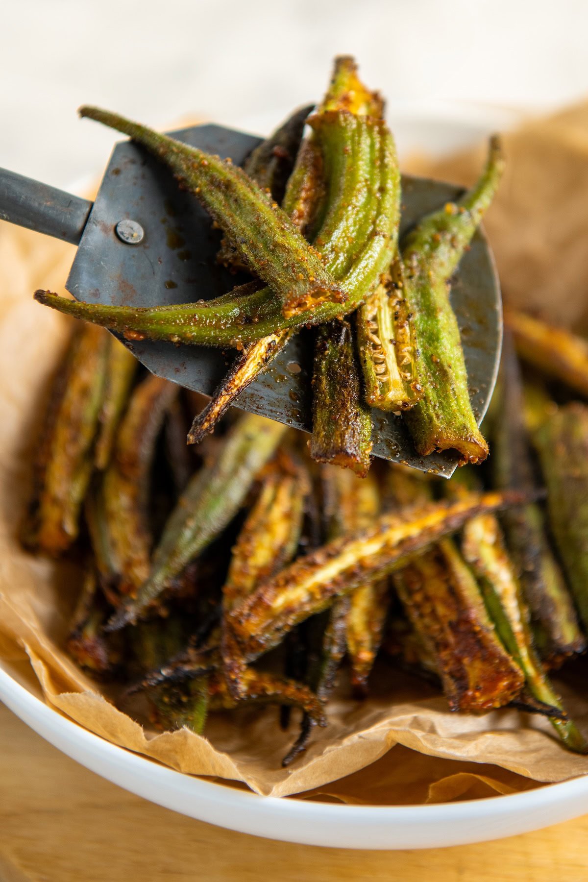 A close-up of a spatula adding air fryer okra into a bowl lined with brown parchment paper.