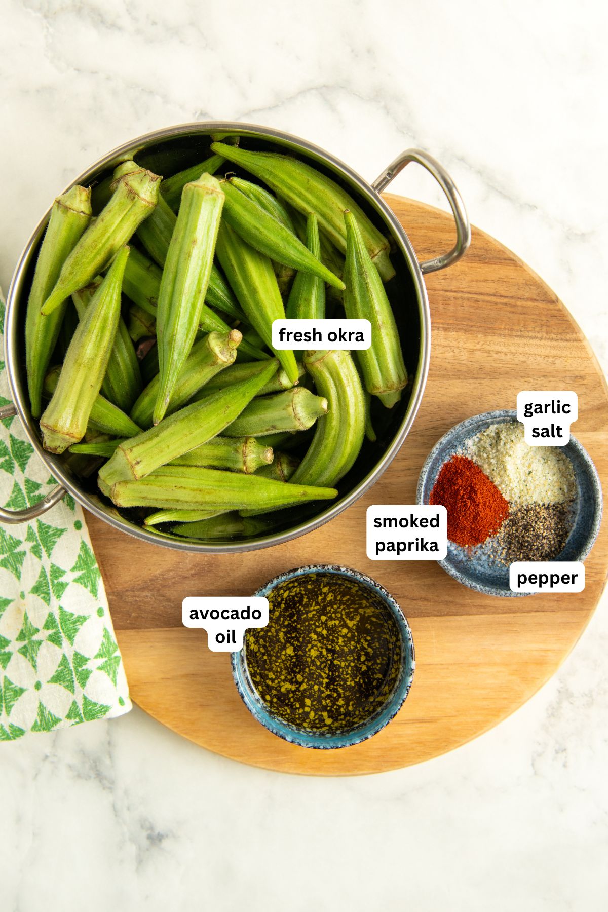 A pot of fresh okra sits on a wooden board, next to a bowl of avocado oil and a small plate with garlic salt, smoked paprika, and pepper.