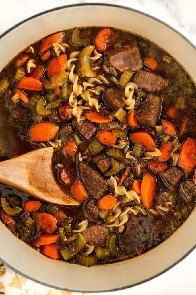 A close-up of a pot filled with beef soup containing chunks of beef, sliced carrots, celery, and spiral noodles in a dark broth. A wooden spoon is partially submerged, stirring the ingredients.