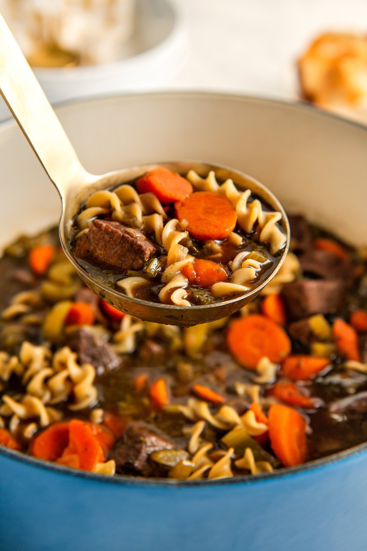 A close-up of a ladle lifting beef and noodle soup with carrots, beef chunks, and spiral pasta from a large pot.