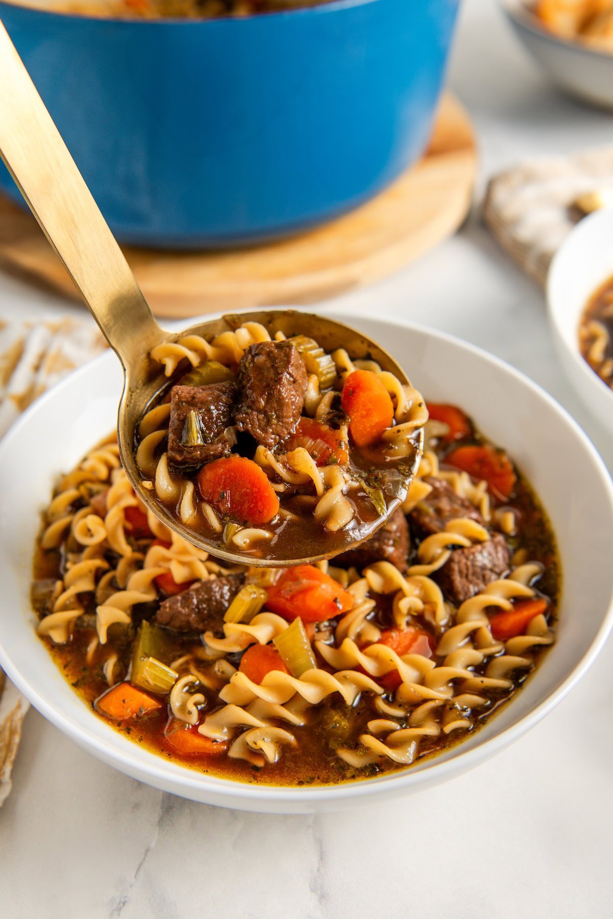 A bowl of beef noodle soup with fusilli pasta, carrots, celery, and chunks of beef, with a ladle lifting a serving above the bowl.